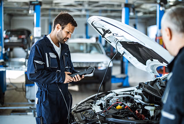 Ein Kfz-Sachverständiger für Gutachten in Berlin und Brandenburg prüft mit einem elektronischen Diagnosegerät den Motorraum eines weißen Autos in einer modernen Werkstatt.